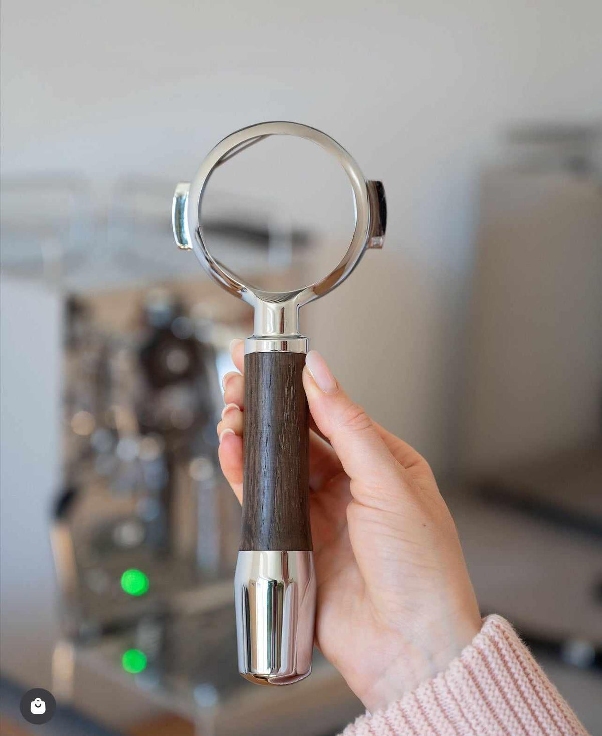 Hand håller ett rostfritt portafilter med mörkt trähandtag framför en kaffemaskin i köket.