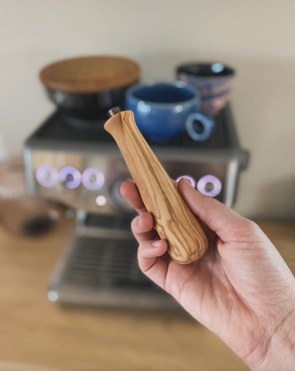 Hand som håller ett portafilterhandtag i trä med espressomaskin och kaffekoppar i bakgrunden.