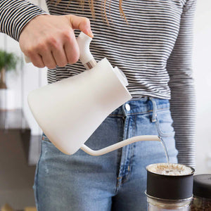 Mão a verter água de um jarro de água branco com bico estreito para um filtro de café, parte do EspressoGear, tudo sobre café.